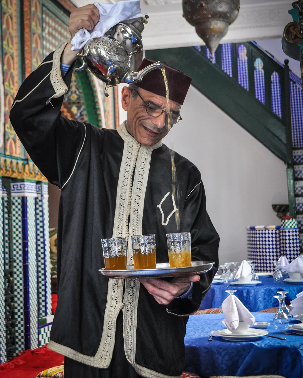 A waiter in a black embroidered robe and fez pours mint tea from a raised silver teapot into small glasses on a tray