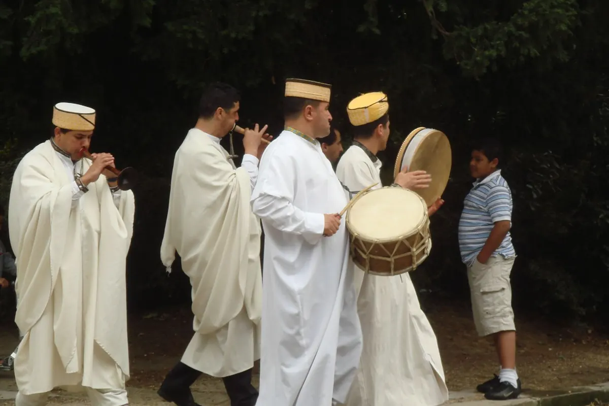 Four men in white robes and caps walk along a pavement playing pipes and a large frame drum in a Moroccan wedding procession